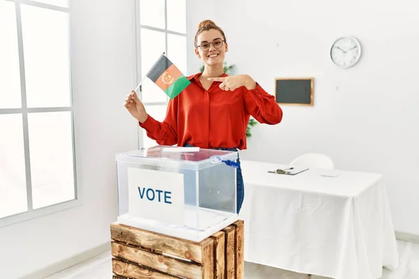 Młoda Brunetka Przez Urnę Głosowania Trzymając Flagę Afganistanu Uśmiechając Się — Zdjęcie stockowe