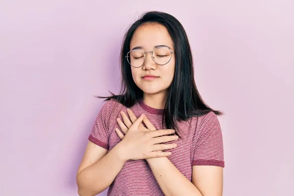 若い中国の女の子は目を閉じて胸の上に手で笑みを浮かべてカジュアルな服や眼鏡を着用し 顔に感謝のジェスチャー 健康の概念 — ストック写真