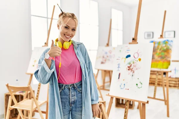 Young caucasian girl at art studio doing happy thumbs up gesture with hand. approving expression looking at the camera showing success.