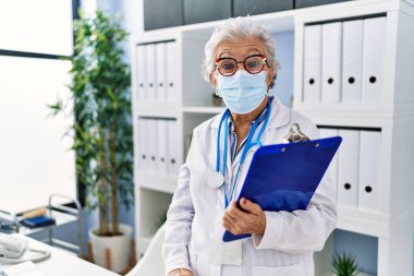 Gri saçlı, doktor üniforması giyen, güvenlik maskesi takan ağzı açık, korkmuş ve şaşırmış yaşlı bir kadın. 