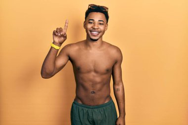 Mayo ve yüzücü gözlüğü takan Afro-Amerikan genç adam bir numarayı gösterip işaret ederken kendinden emin ve mutlu bir şekilde gülümsüyor.. 