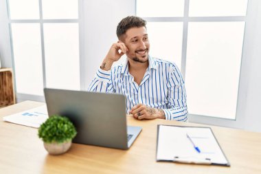 Ofiste çalışan sakallı genç yakışıklı bir adam bilgisayarını kullanıyor. Yüzünde gülümseme ve doğal ifadeyle yan yana bakıyor. Kendinden emin gülen. 