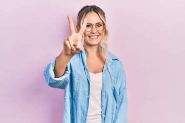 Güzel, genç, sarışın bir kadın. Günlük kıyafetler ve gözlük takıyor. İki numaralı parmaklarıyla gösteriyor ve işaret ediyor.. 