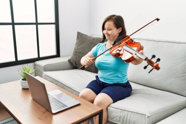 Down sendromlu esmer kadın kanepede oturup oturma odasında keman çalışıyor.
