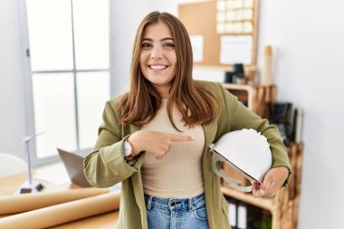 Genç esmer kadın, ofiste mimar şapkası tutuyor, gülümsüyor, mutlu bir şekilde eli ve parmağı ile işaret ediyor. 