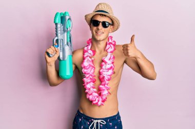 Beyaz mayo giyen genç bir adam ve elinde su tabancası tutan Hawai leyleği mutlu ve pozitif gülümsüyor, başparmağıyla mükemmel bir iş çıkarıyor ve onay işareti yapıyor. 