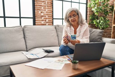 Orta yaşlı kadın kumar paraları sayıyor ve evde dizüstü bilgisayar kullanıyor.