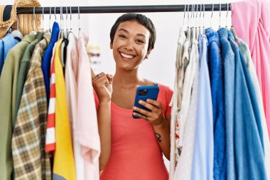 Kısa saçlı İspanyol bir kadın elbise rafında kıyafet arıyor. Akıllı telefon kullanıyor. Gülümsüyor. 
