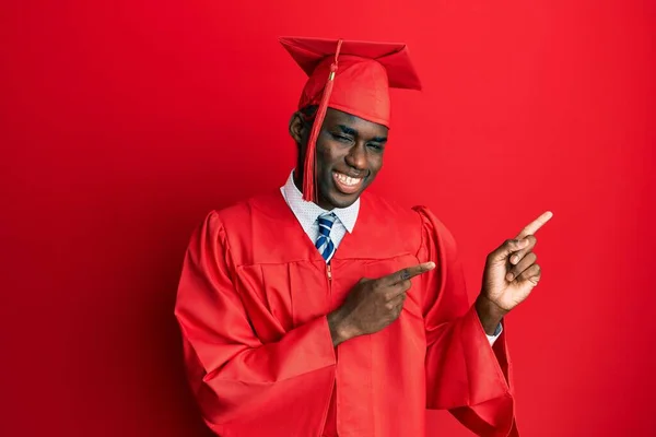 若いアフリカ系アメリカ人の男が卒業キャップと式のローブを着て笑顔とカメラを見て2本の手と指で側面を指して — ストック写真