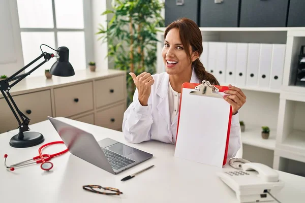 Hispanic Doctor Woman Holding Clipboard Pointing Thumb Side Smiling Happy — Stock Photo, Image