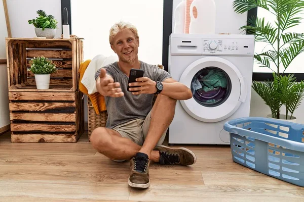Genç Sarışın Adam Çamaşır Yıkıyor Akıllı Telefon Kullanıyor Gülümsüyor Dostça — Stok fotoğraf