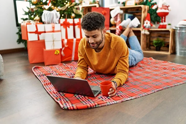 年轻的Arab男子使用笔记本电脑 在家里的圣诞树旁躺在地板上喝咖啡 — 图库照片