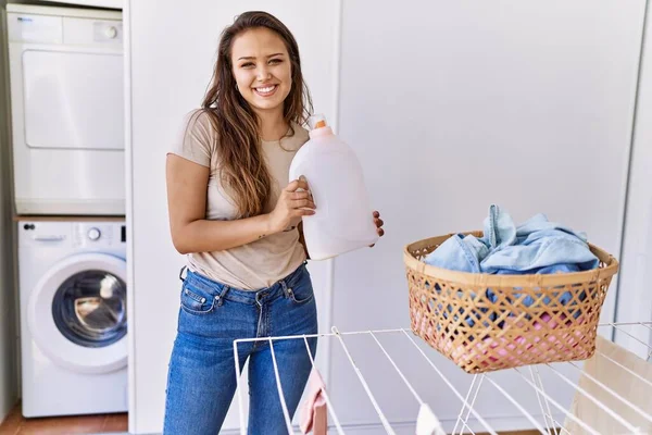 Young hispanic girl doing laundry holding detergent bottle at home