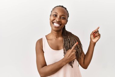 Örgü örgülü siyahi kadın izole edilmiş arka planda duruyor gülümsüyor ve kameraya bakıyor iki eliyle ve parmaklarıyla kenarı işaret ediyor.. 