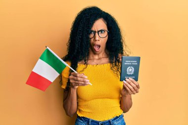 Orta yaşlı Afro-Amerikalı kadın elinde İtalyan bayrağı ve pasaportuyla şaşkın, şüpheci ve alaycı görünüyor, ağzı açık kalmış ve şaşırmış. 