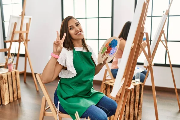 若いヒスパニックアーティストの女性がアートスタジオでキャンバスに絵を描くのは 勝利のサインをしている指を示すカメラを見て笑っています — ストック写真