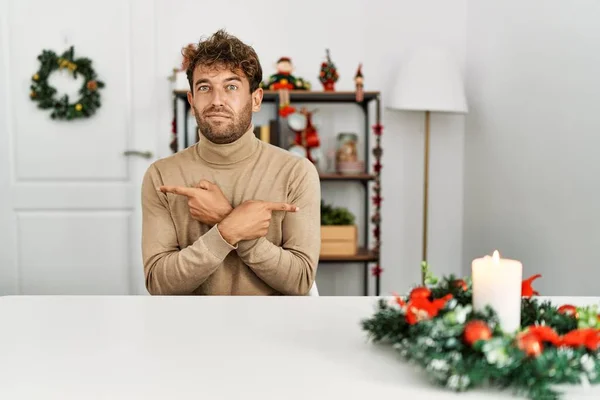 若いですハンサムな男でひげがテーブルの上に座ってクリスマスの装飾指で両側を指して 異なる方向が反対 — ストック写真