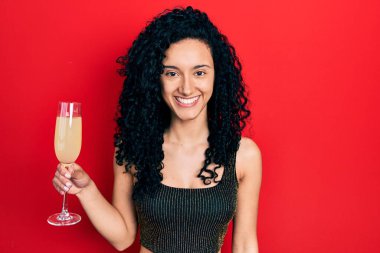 Kıvırcık saçlı genç İspanyol kadın bir bardak şampanya içiyor. Pozitif ve mutlu görünüyor. Kendine güvenen bir gülümsemeyle dişlerini gösteriyor. 