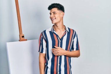 Genç İspanyol bir adam boş tuvale yakın duruyor. Yüzünde gülümseme, doğal bir ifade ile yan yana bakıyor. Kendinden emin gülen. 