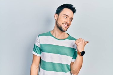 Gündelik kıyafetler giyen, gülümseyen, gülümseyen ve başparmağıyla yan tarafı gösteren İspanyol genç adam.. 