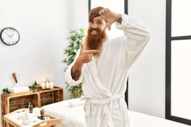 Uzun sakallı, sağlıklı spa 'da bornoz giyen, gülümseyen, elleri ve parmakları mutlu yüzlü kızıl saçlı bir adam. yaratıcılık ve fotoğrafçılık kavramı. 