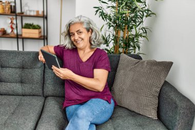 Orta yaşlı, kır saçlı, dokunmatik ped kullanan kadın evdeki koltukta oturuyor..