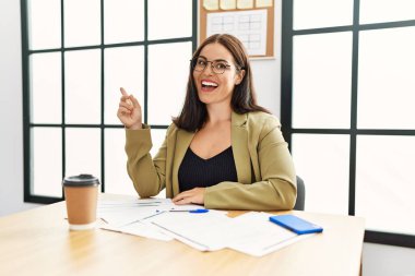 İş stili giyinmiş genç esmer kadın ofiste oturmuş gülümsüyor mutlu bir şekilde el ve parmak kenarı işaret ediyor. 