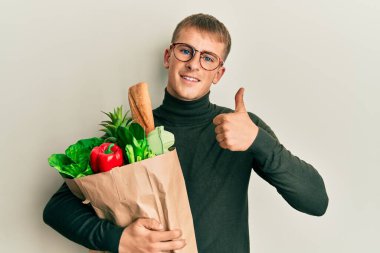 Elinde alışveriş torbalarıyla gülümseyen beyaz bir genç, mutlu ve pozitif, başparmağıyla mükemmel ve onay işareti yapıyor. 