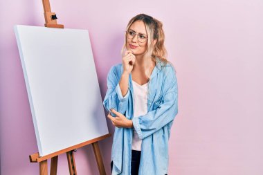 Güzel sarışın kadın beyaz boyacı sehpası başında durup parmak çenesine değmiş şüpheyi düşünüp kafasını kaldırıyor. 