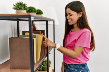 Genç Latin kadın gülümsüyor. Kendine güveni tam. Evde raflık kitap seçiyor.