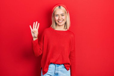 Güzel sarışın kadın kırmızı süveter giyer ve parmaklarıyla dört numarayı gösterirken kendine güvenen ve mutlu bir şekilde gülümser.. 