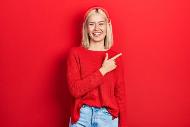 Güzel sarışın kadın, günlük kırmızı süveter giyiyor, neşeli, yüzünde mutlu ve doğal bir ifadeyle, elleri ve parmaklarıyla işaret eden bir gülümseme var. 