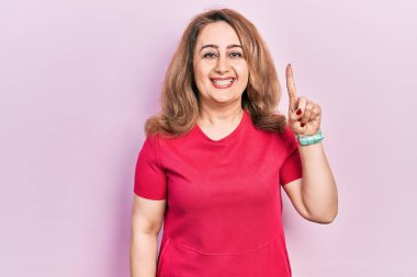 Orta yaşlı beyaz kadın, günlük kıyafetler giyiyor. Bir numarayı gösteriyor ve işaret ediyor. Kendinden emin ve mutlu bir şekilde gülümsüyor.. 