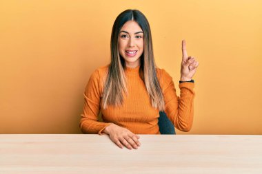 Genç İspanyol kadın gündelik giysiler içinde masada oturuyor ve bir numaralı parmağını gösterip gülümserken kendinden emin ve mutlu. 