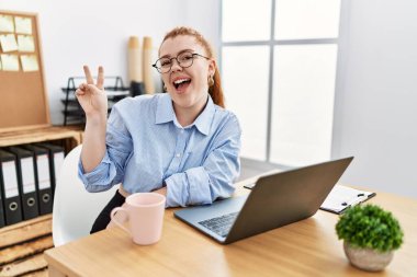 Ofiste çalışan genç kızıl saçlı kadın bilgisayarlı dizüstü bilgisayar kullanıyor gülümsüyor ve parmaklarını zafer işareti yaparken kameraya bakıyor. İki numara.. 