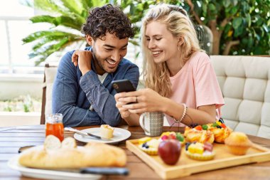 Genç çift kahvaltı yapıyor ve akıllı telefon kullanıyor terasta masada oturuyor..