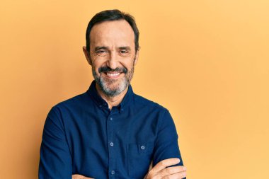 Orta yaşlı İspanyol bir adam sıradan kıyafetler giyip kollarını kavuşturup kameraya bakarken gülümsüyor. pozitif kişi. 