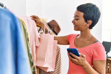 Genç İspanyol kadın müşteri elbise mağazasında akıllı telefon kullanıyor.