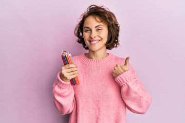 Genç esmer kadın elinde renkli kalemlerle mutlu ve pozitif gülümsüyor, başparmağıyla mükemmel ve onay işareti yapıyor. 