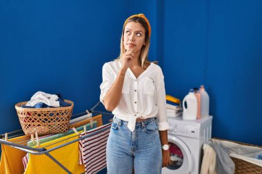 Çamaşırhanede eli çenesinde soru soran genç sarışın bir kadın, düşünceli bir ifade. Düşünceli bir yüzle gülümsüyor. şüphe kavramı. 