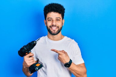 Sakallı genç Arap adam, matkap kullanıyor. Gülümsüyor, el ve parmağıyla işaret ediyor. 