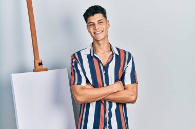 Boş tuvalin yanında duran genç İspanyol adam kollarını kavuşturup kameraya bakarken gülümsüyor. pozitif kişi. 