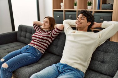 Mutlu bir şekilde gülümseyen genç çift ellerini başının üstüne koyup evdeki kanepede oturuyor..