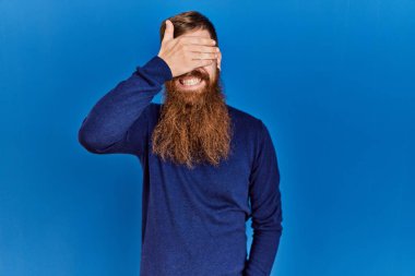 Uzun sakallı kızıl saçlı adam mavi arka planda günlük mavi süveter giyiyor gülümsüyor ve elleri yüzünde şaşkınlıktan gözleri kapayarak gülüyor. kör kavram. 