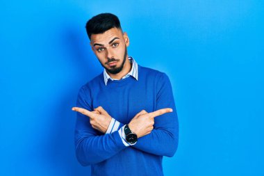 Sakallı, mavi süveterli, iki tarafı da parmaklarıyla gösteren genç İspanyol adam farklı yönlere bakıyor. 