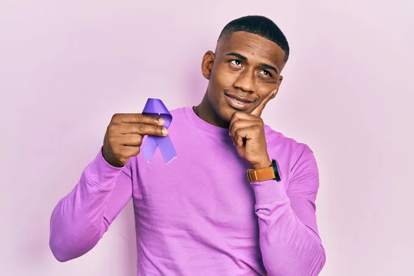 Young black man holding purple ribbon awareness serious face thinking about question with hand on chin, thoughtful about confusing idea