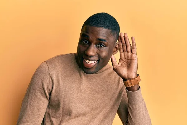 Young african american man wearing casual winter sweater smiling with hand over ear listening and hearing to rumor or gossip. deafness concept.
