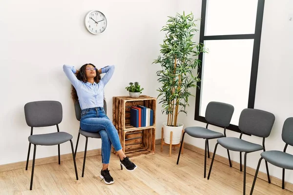 Genç Latin kadın ellerini başının üstüne koyup bekleme odasında müzik dinliyor.