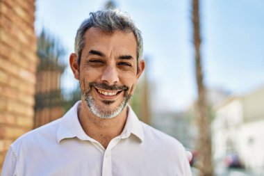 Orta yaşlı, kır saçlı, gülümseyen bir adam şehirde mutlu bir şekilde duruyor..