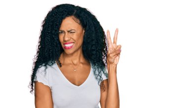 Orta yaşlı Afro-Amerikalı kadın, günlük beyaz tişört giyip kameraya gülümseyen ve zafer işareti yapan. İki numara.. 